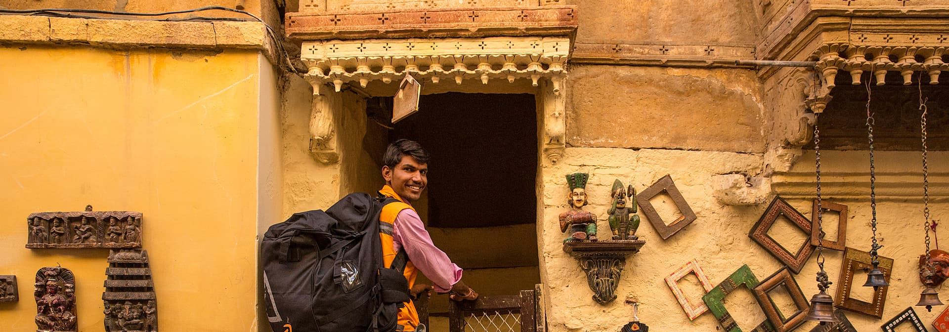An Amazon delivery associate in an orange vest smiles in front of a customer's home in Jaisalmer, surrounded by local crafts and art
