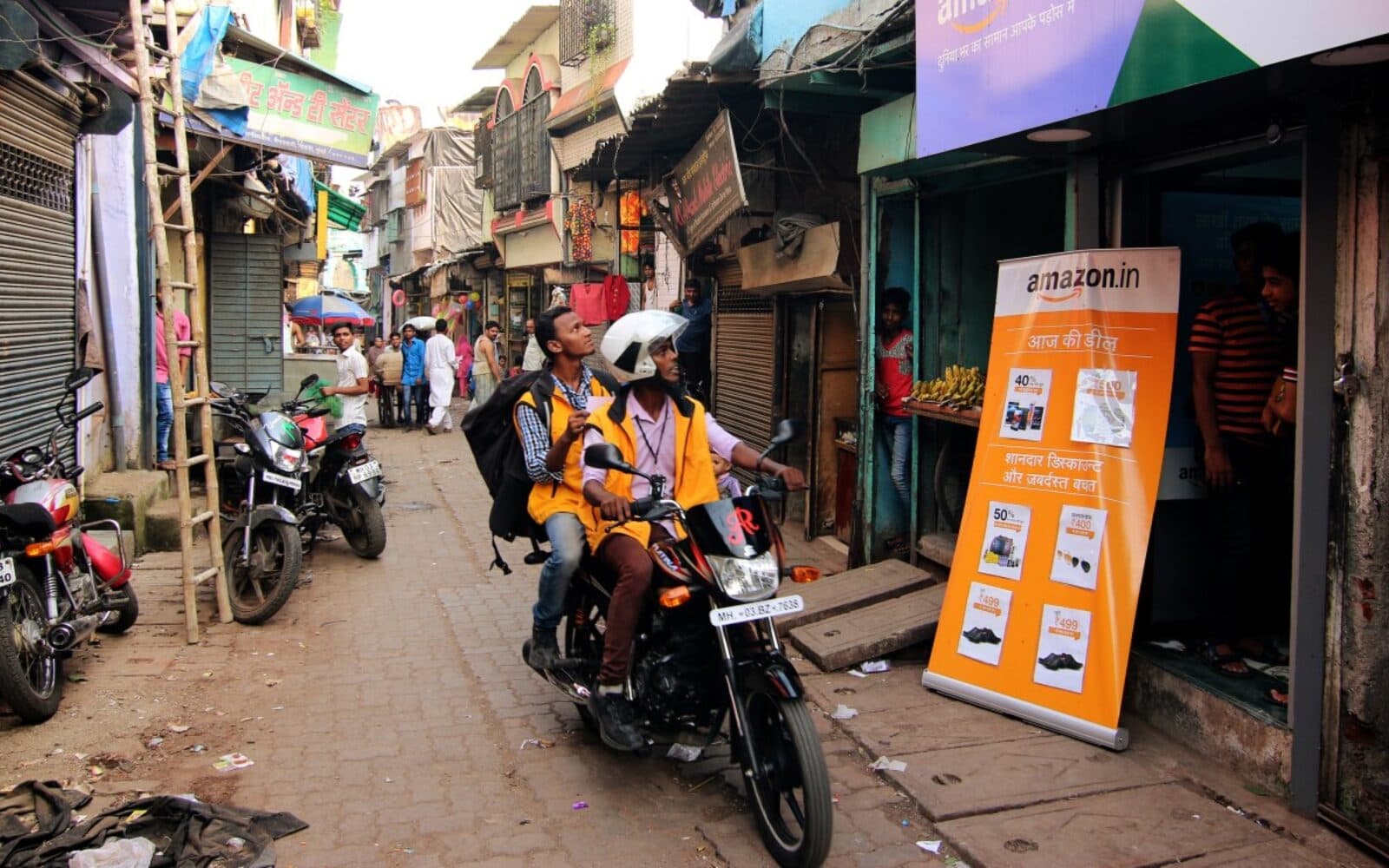 Amazon India delivery associates on a motorcycle on a busy Indian street, passing an amazon.in storefront banner with local-language ads