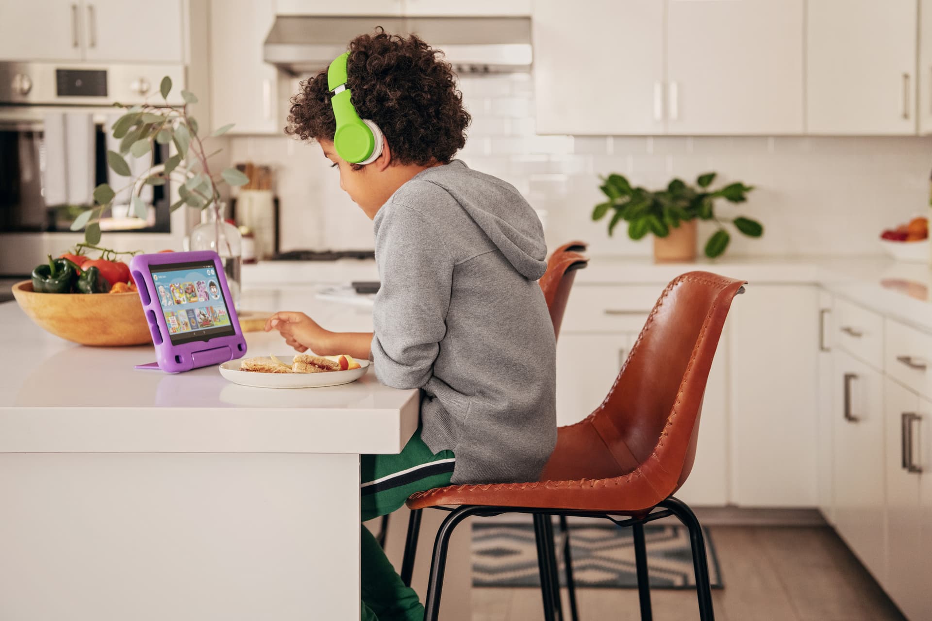 Child with green headphones watching Amazon Kids on a purple Fire HD 8 Kids Edition tablet at a kitchen counter