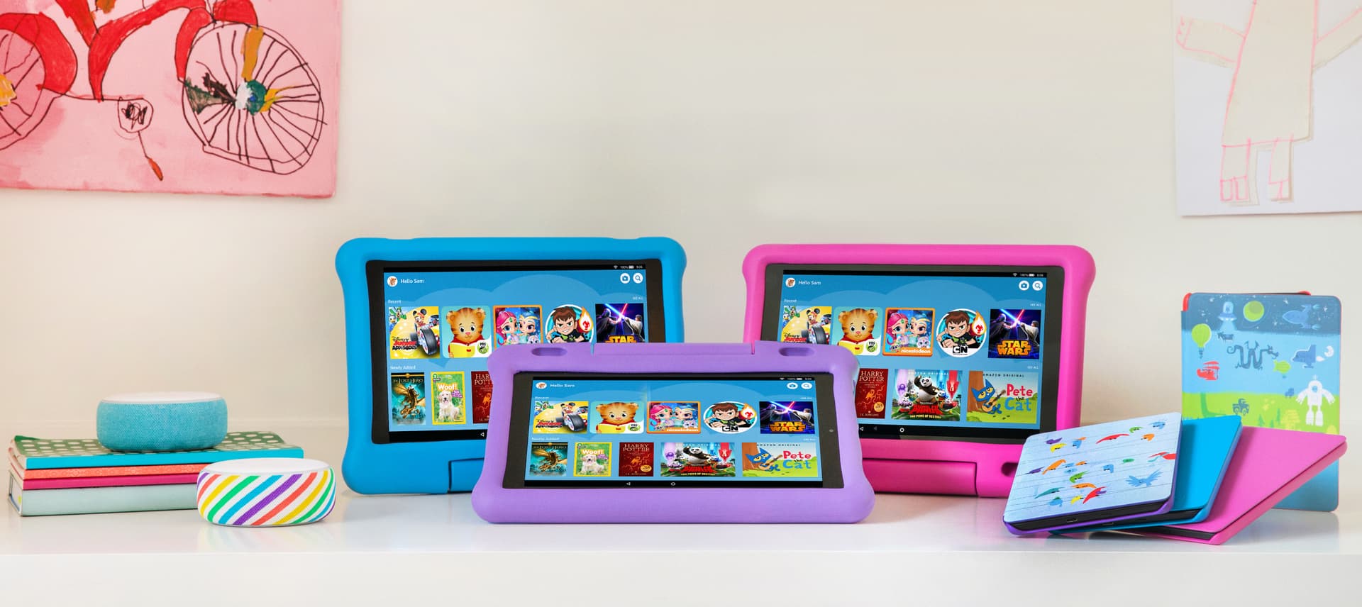 Three Fire Kids Edition tablets in blue, red, and purple cases displaying the Amazon Kids home screen side by side with illustrated Kindle Kids cases