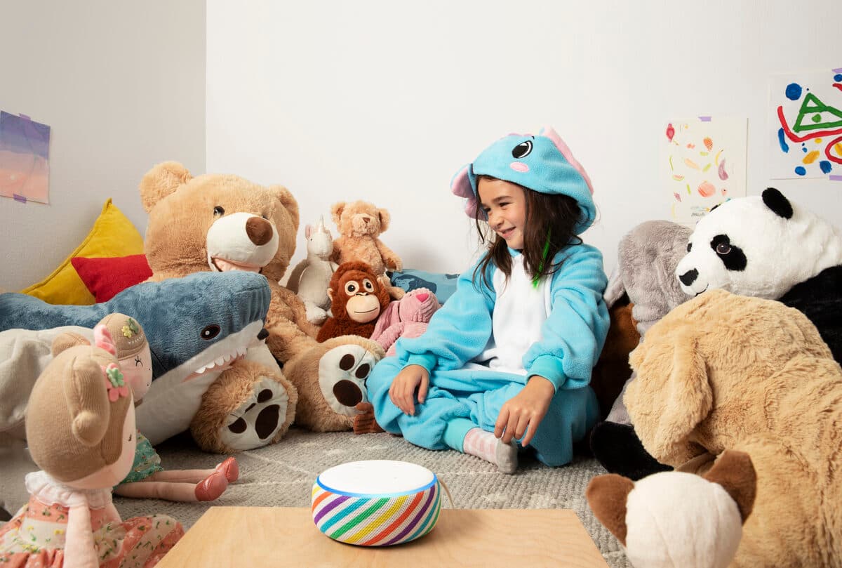 A child in a onesie sits among plush toys with a Rainbow Echo Dot Kids Edition on the table